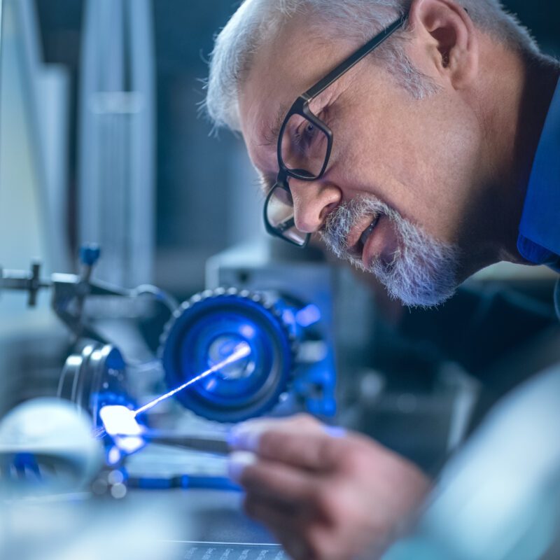 Close-up Portrait of Focused Middle Aged Engineer in Glasses Working with High Precision Laser Equipment, Using Lenses and Testing Optics for Accuracy Required Electronics